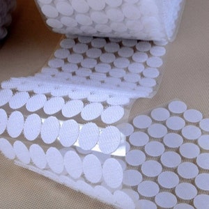 Könnte beinhalten: Eine Rolle weiße Klebepunkte mit durchsichtiger Rückseite. Die Punkte sind in Reihen angeordnet und zum Befestigen von Gegenständen an Oberflächen gedacht.