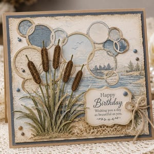 Puede incluir: Una tarjeta de felicitación cuadrada con una ilustración de un paisaje lacustre con espadañas en primer plano. La tarjeta incluye recortes circulares superpuestos, pequeñas cuentas azules y una etiqueta con el texto Happy Birthday Wishing you a day as beautiful as you.
