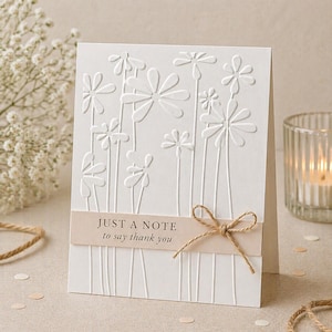 May include: A white greeting card with embossed floral design. The card features the text "JUST A NOTE to say thank you" on a beige band, tied with a twine bow. The card is set on a neutral surface.