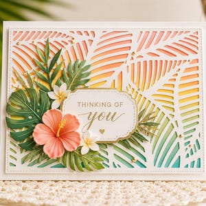 Pode incluir: Um cartão branco com tema tropical. O cartão apresenta um design cortado a laser com um gradiente de laranja, amarelo e verde. Enfeitado com flores de papel, folhas e o texto "Thinking of You".