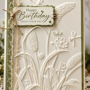 May include: A cream-colored greeting card featuring embossed white botanical designs of grasses, flowers, a ladybug, and dragonflies. A rectangular tag with the text Happy Birthday ENJOY YOUR DAY is attached with twine, accented by a strip of burlap and green paper.