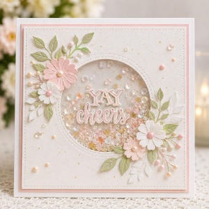 Può includere: Biglietto di auguri quadrato con sfondo bianco e bordo rosa. La carta presenta una finestra circolare piena di coriandoli e le parole "YAY cheers". Fiori rosa e bianchi e foglie verdi decorano la carta.