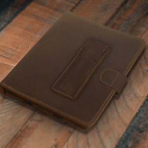 May include: A brown leather journal with a pen holder and a snap closure. The journal is rectangular and has a textured surface. The pen holder is attached to the front of the journal with white stitching.