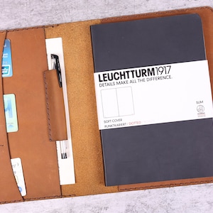 May include: A brown leather notebook cover with a pen holder and multiple card slots. The cover is open to reveal a black notebook with the text "LEUCHTTURM1917" and "DETAILS MAKE ALL THE DIFFERENCE." The notebook is a soft cover with dotted pages.