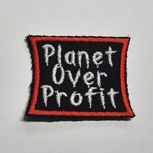 May include: Embroidered patch with the words "Planet Over Profit" in white script on a black background. The patch has a red border and is square-shaped. The patch is likely for clothing or accessories.