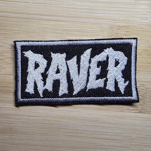 Op de afbeelding: Een geborduurde zwarte en witte patch met het woord "RAVER" in witte letters op een zwarte achtergrond.
