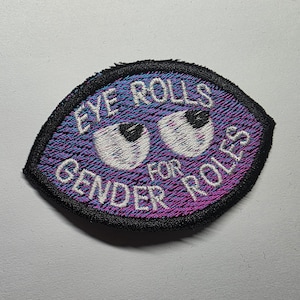 Puede incluir: Parche bordado con forma de ojo, con las palabras "EYE ROLLS FOR GENDER ROLES". El parche tiene un fondo degradado en azul, morado y rosa, con un borde negro. El diseño del ojo presenta dos globos oculares blancos con pupilas negras.