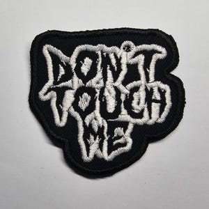 May include: Black embroidered patch with the words "DON'T TOUCH ME" in white, stylized lettering. The patch has a black border and is likely designed to be sewn or ironed onto clothing or accessories.
