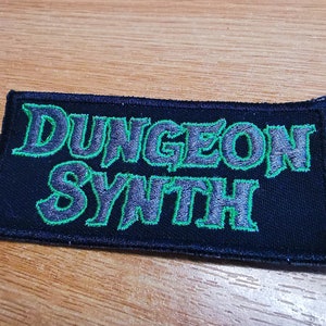 May include: A black fabric patch with green embroidered text that reads "Dungeon Synth".