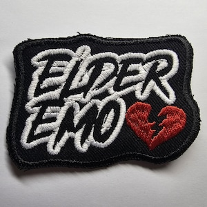 May include: Black embroidered patch with the words "ELDER EMO" in white script. A red, broken heart is on the right side. The patch has a black border and is likely for clothing or accessories.