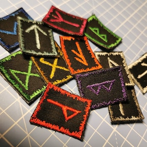 May include: A collection of 12 black fabric patches with different embroidered runes in various colours, including white, green, yellow, orange, purple, and light blue. The patches are arranged on a blue gridded surface.