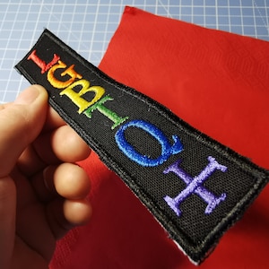 Könnte beinhalten: Schwarzer rechteckiger Aufnäher mit dem Wort "LGBTQ" in Regenbogenfarben gestickt. Der Aufnäher wird vor einem roten Hintergrund gehalten. Die Buchstaben sind in einer serifenlosen Schriftart gehalten.