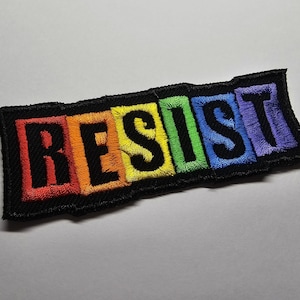 May include: Embroidered patch with the word "RESIST" in black capital letters. The letters are set against a rainbow gradient background, with each letter in a different color. The patch has a black border.