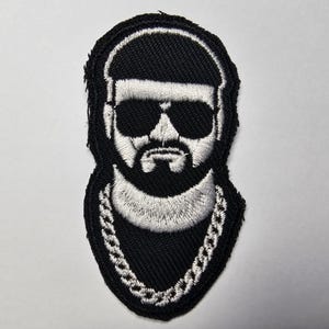 Pode incluir: Patch bordado com um retrato estilizado de um homem com óculos de sol, barba e colar de corrente. O design é principalmente em preto e branco, com os detalhes do rosto e da corrente em fio branco sobre um fundo preto.