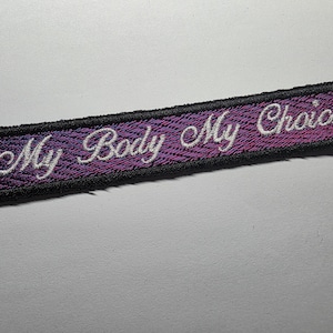 May include: A rectangular embroidered patch with the words "My Body My Choice" in white script. The background is a purple and pink gradient, framed by a black border.
