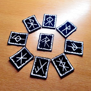 May include: Ten black square patches with white embroidered runes arranged in a circle on a wooden surface.
