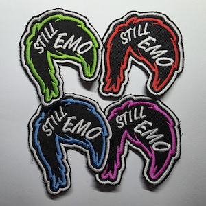 May include: Four embroidered patches with the text "STILL EMO" in white. Each patch is a different colour: green, red, blue, and purple, with a black background and white border. The patches have a unique, stylized shape.