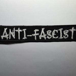 May include: Black rectangular patch with the embroidered text "ANTI-FASCIST" in white. The patch is likely made of fabric and features a stitched border. The text is in a bold, sans-serif font.