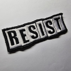Puede incluir: Parche bordado con la palabra "RESIST" en mayúsculas negras. Las letras están rellenas de hilo blanco y delineadas con un borde negro. El parche tiene forma rectangular con un borde ligeramente irregular.
