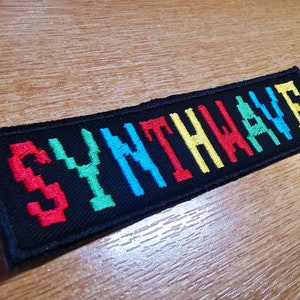 Puede incluir: Un parche de tela negro con la palabra "SYNTHWAVE" bordada en letras pixeladas rojas, verdes, azules y amarillas.