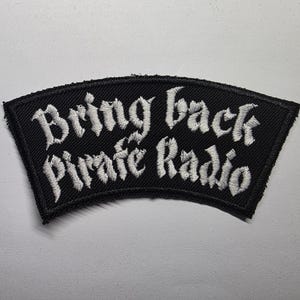 May include: Black, arched patch with white embroidered text reading "Bring back Pirate Radio." The patch has a black border and is likely designed to be sewn onto clothing or accessories. The text is in a gothic font.