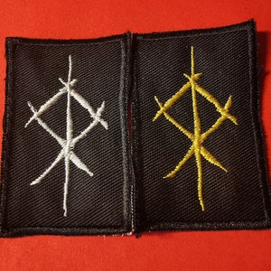 May include: Two black fabric patches with embroidered white and gold symbols. The symbols are similar in design, but one is white and the other is gold.