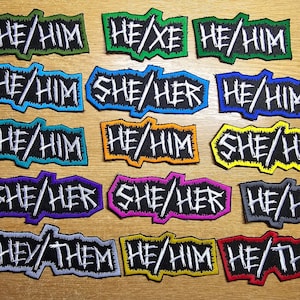 May include: A collection of 15 colourful iron-on patches with different pronoun combinations, including HE/HIM, SHE/HER, THEY/THEM, and HE/XE. The patches are all black with white lettering and have a rough, hand-drawn style.