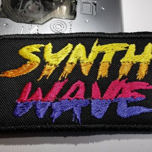 Puede incluir: Un parche bordado negro con el texto "Synth Wave" en amarillo, naranja, rosa y morado.