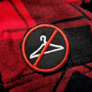 May include: A red and black plaid fabric with a black and white embroidered patch. The patch is a circle with a red line through it and a white hanger inside. The text "No Hangers" is not visible in the image.