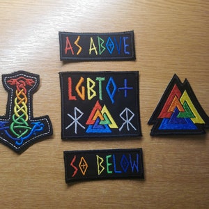 Op de afbeelding: Vijf geborduurde patches met regenboogkleuren en zwarte achtergronden. De patches tonen een hamer, een driehoek en de tekst "LGBTQ+ RAWR", "AS ABOVE" en "SO BELOW".
