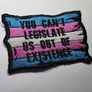 Può includere: Una toppa rettangolare ricamata con strisce orizzontali azzurre, rosa e bianche. La toppa presenta un bordo nero cucito e riporta il testo ricamato in nero: YOU CAN'T LEGISLATE US OUT OF EXISTENCE al centro.