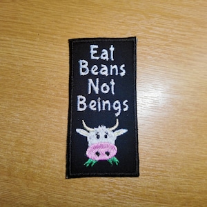 Könnte beinhalten: Schwarzer gestickter Aufnäher mit weißem Text, der "Eat Beans Not Beings" lautet, und einer Comic-Kuh mit rosa Nase und grünem Gras im Maul.