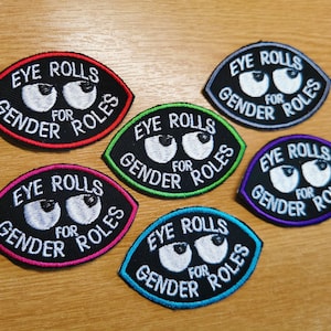 Könnte beinhalten: Sechs gestickte Aufnäher mit dem Text "Eye Rolls for Gender Roles" in weißer Schrift auf schwarzem Hintergrund. Die Aufnäher sind in verschiedenen Farben: rot, grün, lila, pink, blau und grau.