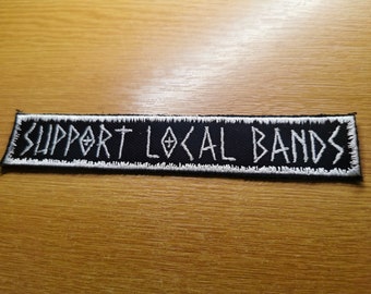 Puede incluir: Parche bordado en blanco y negro con el texto "SUPPORT LOCAL BANDS" en una fuente estilizada.