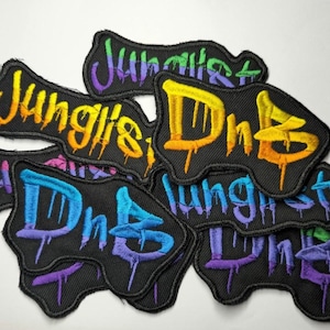 May include: Several black embroidered patches featuring the words "Junglist DnB" in a graffiti style. The lettering is in bright colours including yellow, orange, blue, and purple, with dripping paint effects.