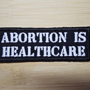 May include: Black rectangular patch with white embroidered text that reads "ABORTION IS HEALTHCARE". The patch has a black border and is set against a light brown wooden surface.