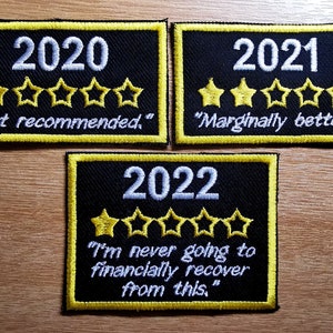 以下が含まれることがあります： 年と星評価がついた、黒と黄色の刺繍パッチが3つ。2020年のパッチは星が1つで、「おすすめしません」と書いてあります。2021年のパッチは星が2つで、「少しはまし」と書いてあります。2022年のパッチは星がなく、「これからはもう経済的に立ち直れない」と書いてあります。