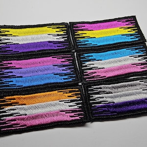 May include: Six embroidered patches with the colors of the genderqueer pride flag. The patches are black with white stitching and feature a design of jagged lines.