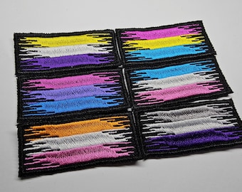 May include: Six embroidered patches with the colors of the genderqueer pride flag. The patches are black with white stitching and feature a design of jagged lines.