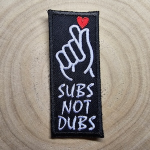 Könnte beinhalten: Schwarzer gestickter Patch mit einer weißen Handgeste und dem Text "SUBS NOT DUBS" in weiß. Ein kleines rotes Herz befindet sich über der Hand.