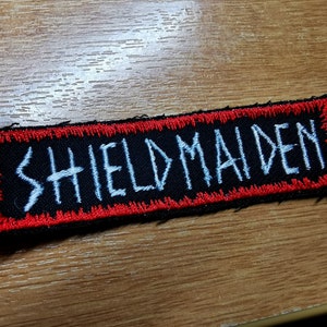 Może przedstawiać: Czarna łatka z tkaniny z czerwoną obwódką. Łatka ma wyszyty biały tekst, który brzmi "SHIELDMAIDEN".