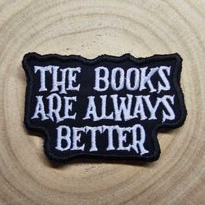 以下が含まれることがあります： 白い文字で「THE BOOKS ARE ALWAYS BETTER」と書かれた黒い刺繍パッチ。 パッチは黒い縁取りがあり、木目調の背景に配置されています。 文学をテーマにしたアクセサリー。