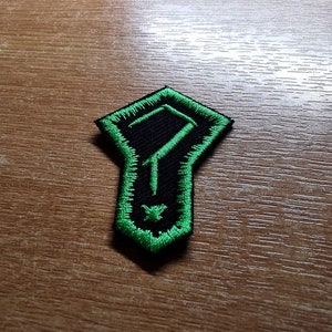 Może przedstawiać: Naszywka haftowana w kolorze czarnym i zielonym z  stylizowanym symbolem. Symbol to stylizowana litera "F" z krzyżem na dole.