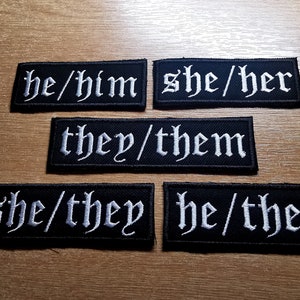 Op de afbeelding: Vijf zwarte stoffen patches met witte geborduurde tekst. De tekst op de patches luidt "he/him", "she/her", "they/them", "she/they" en "he/they".