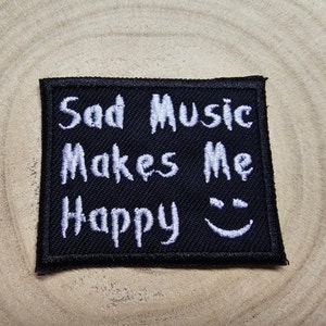 May include: A black embroidered patch with the words "Sad Music Makes Me Happy" in white, and a smiling face. The patch has a black border and is displayed on a wooden surface.