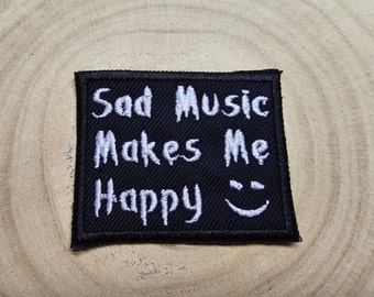 Pode incluir: Um patch bordado preto com as palavras "Sad Music Makes Me Happy" em branco, juntamente com uma carinha sorridente. O patch tem uma borda preta e é exibido em uma superfície de madeira.