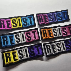 Könnte beinhalten: Gestickte Aufnäher mit dem Wort "RESIST" in verschiedenen Farbkombinationen. Jeder Aufnäher hat einen schwarzen Rand und das Wort ist in leuchtenden Farben wie Pink, Blau, Gelb und Lila gestickt. Die Aufnäher sind rechteckig.