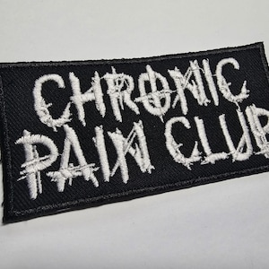 Könnte beinhalten: Schwarzer rechteckiger Aufnäher mit weiß gesticktem Text „CHRONIC PAIN CLUB“. Der Aufnäher hat einen schwarzen Rand und ist wahrscheinlich aus Stoff. Der Text hat eine verwegene, kantige Schriftart.