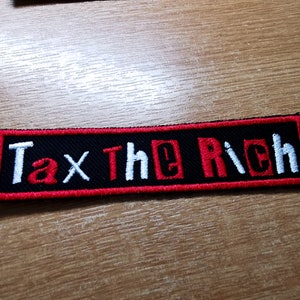 Könnte beinhalten: Ein gestickter schwarzer und roter Aufnäher mit dem Text "Tax The Rich" in weißen Buchstaben.