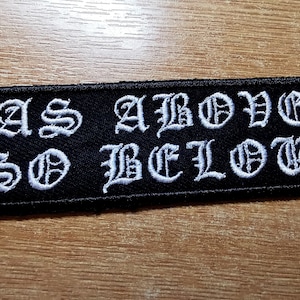 May include: A black rectangular patch featuring the embroidered phrase "AS ABOVE SO BELOW" in white gothic script. This patch, suitable for clothing or bags, has a dark, gothic style.